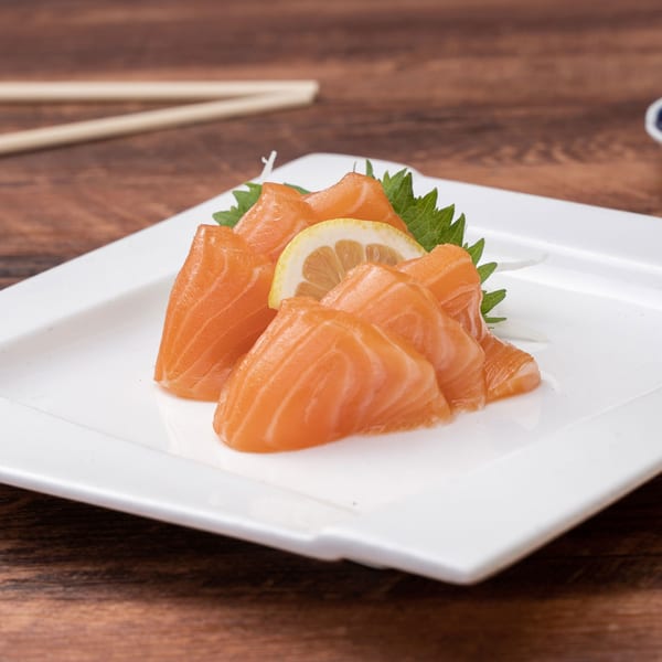 Fresh salmon sashimi slices with a lemon wedge and green garnish on a white plate.