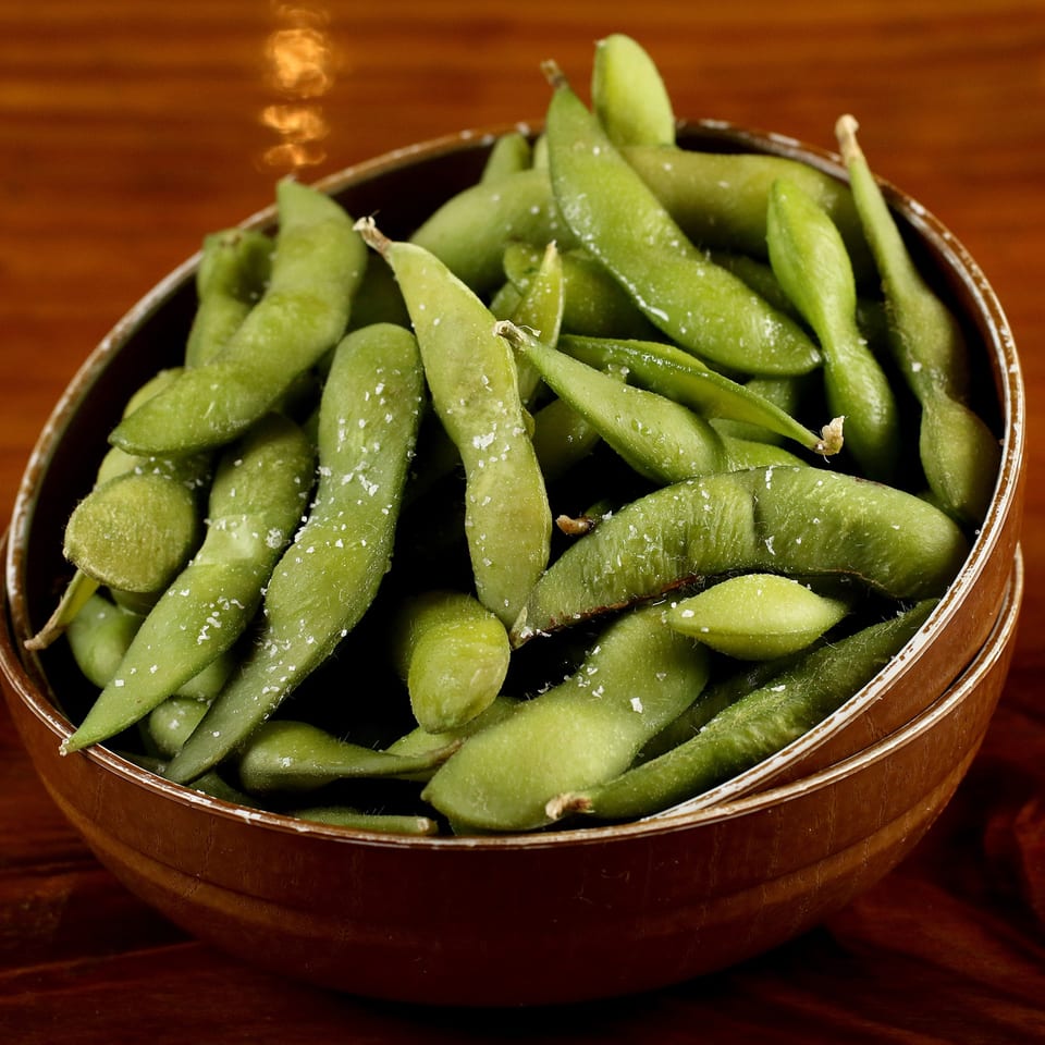 A brown bowl filled with bright green, salted edamame pods on a dark wooden surface.