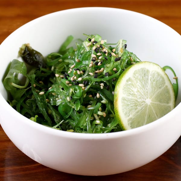 A white bowl filled with green seaweed salad, topped with sesame seeds and a lime slice.